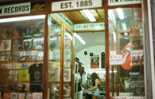 Fachada de uma loja de discos de vinil, vendo-se no interior uma rapariga pegando num disco