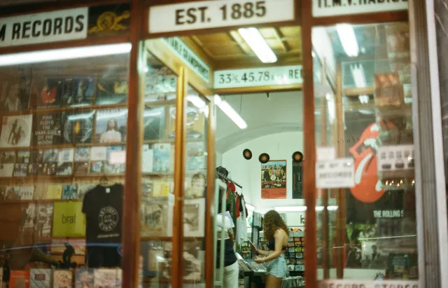 Fachada de uma loja de discos de vinil, vendo-se no interior uma rapariga pegando num disco